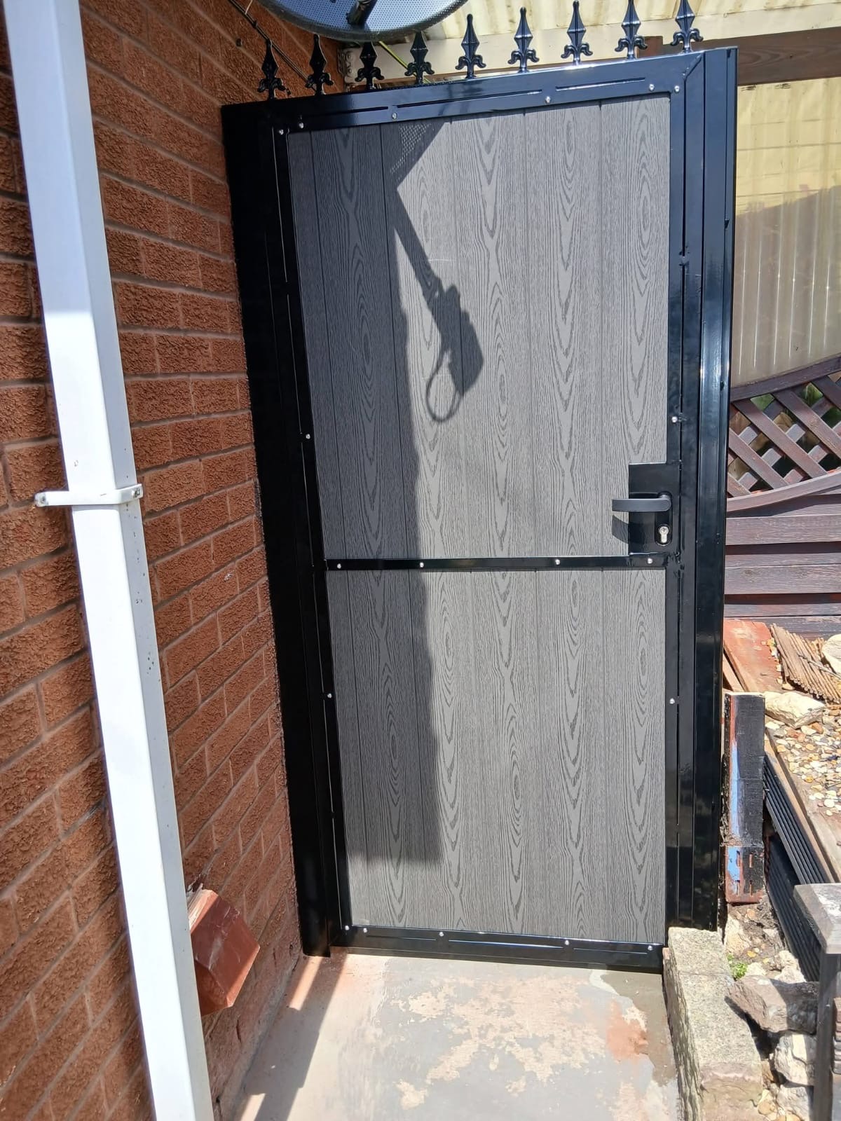 Grey composite pedestrian gate with black metal frame and decorative finials between brick walls