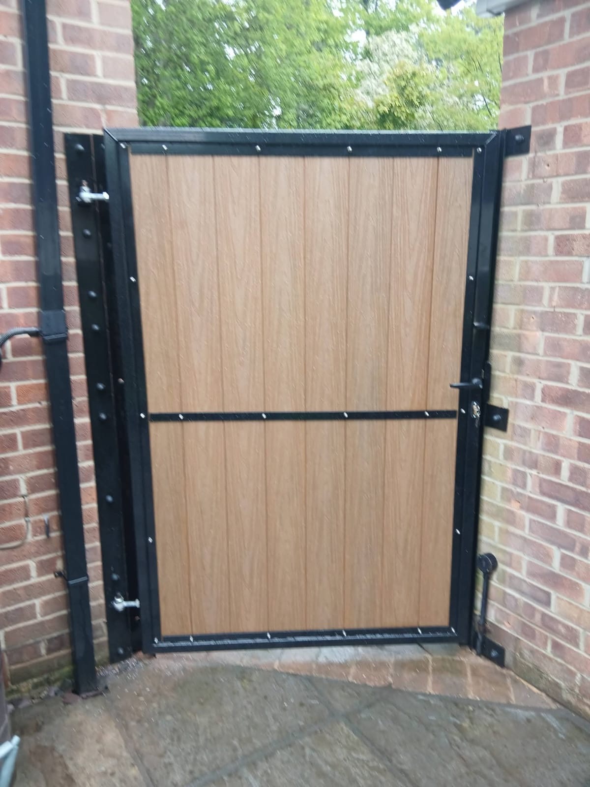 Golden oak effect composite pedestrian gate with black metal frame between red brick walls