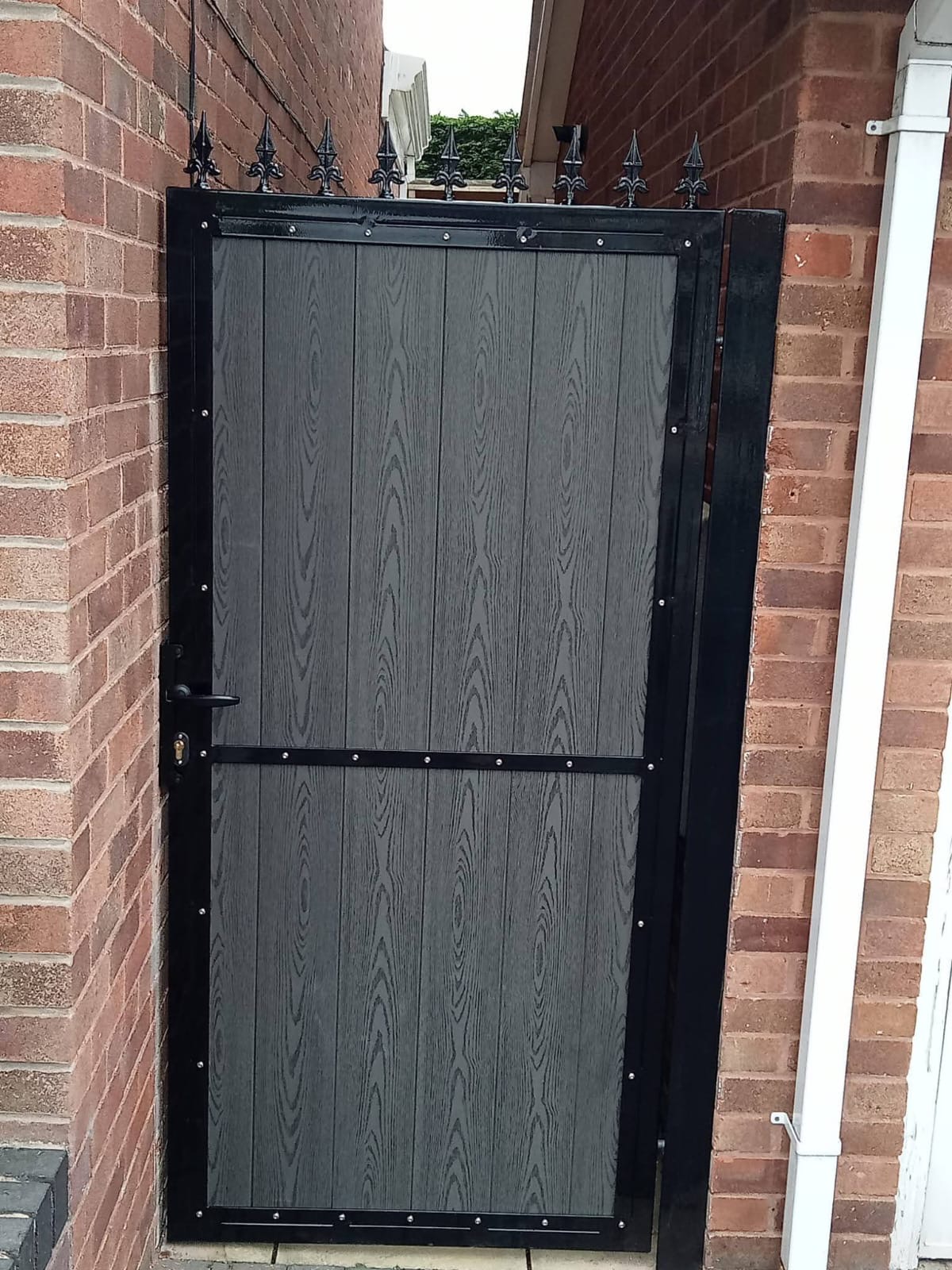Grey wood-effect composite side gate with spear-top metal frame in a narrow brick passageway