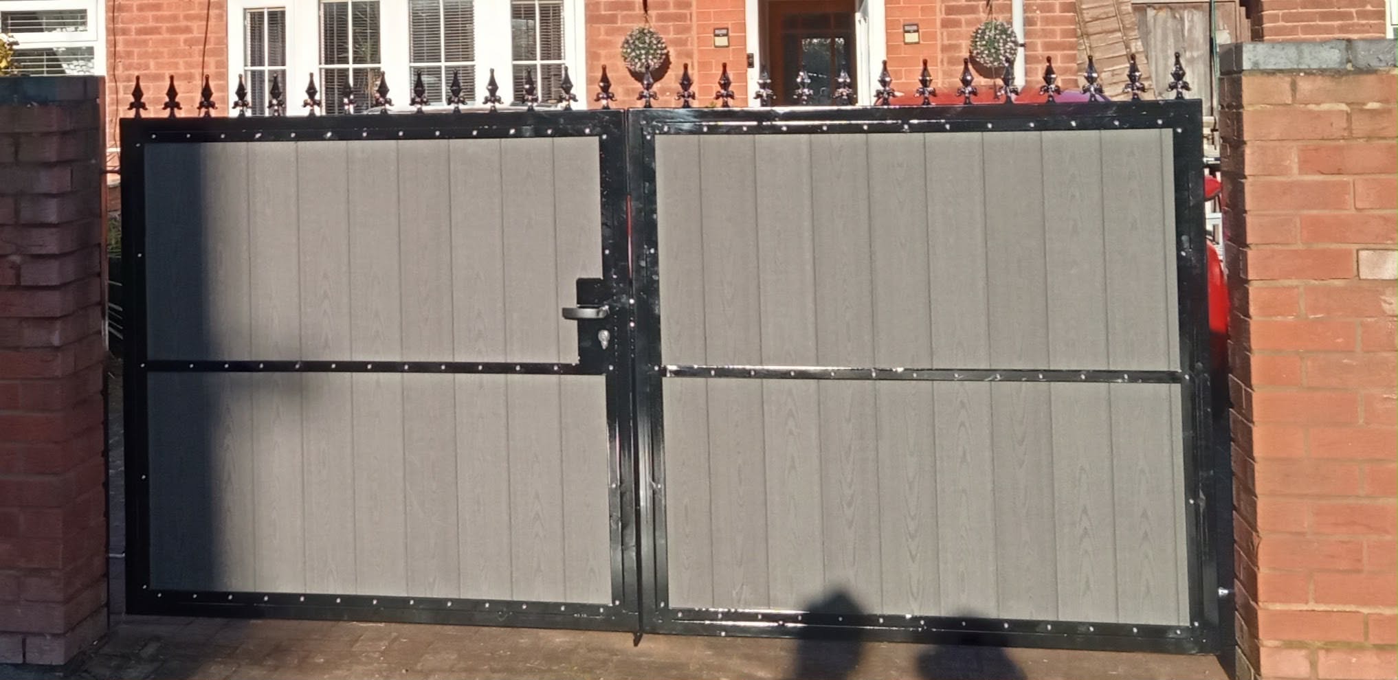 Double composite driveway gates with grey wood-effect panels and black metal frame between brick pillars